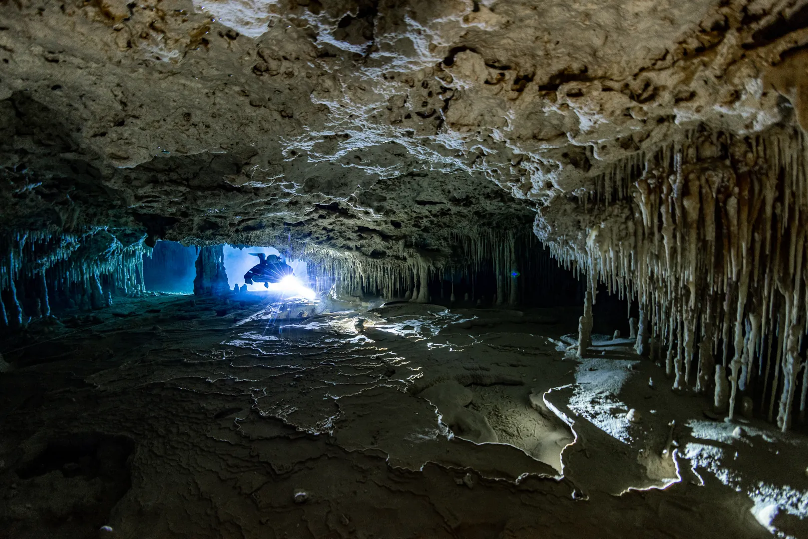 Technical diver swimming through a dark underwater cave with bright lights illuminating formations